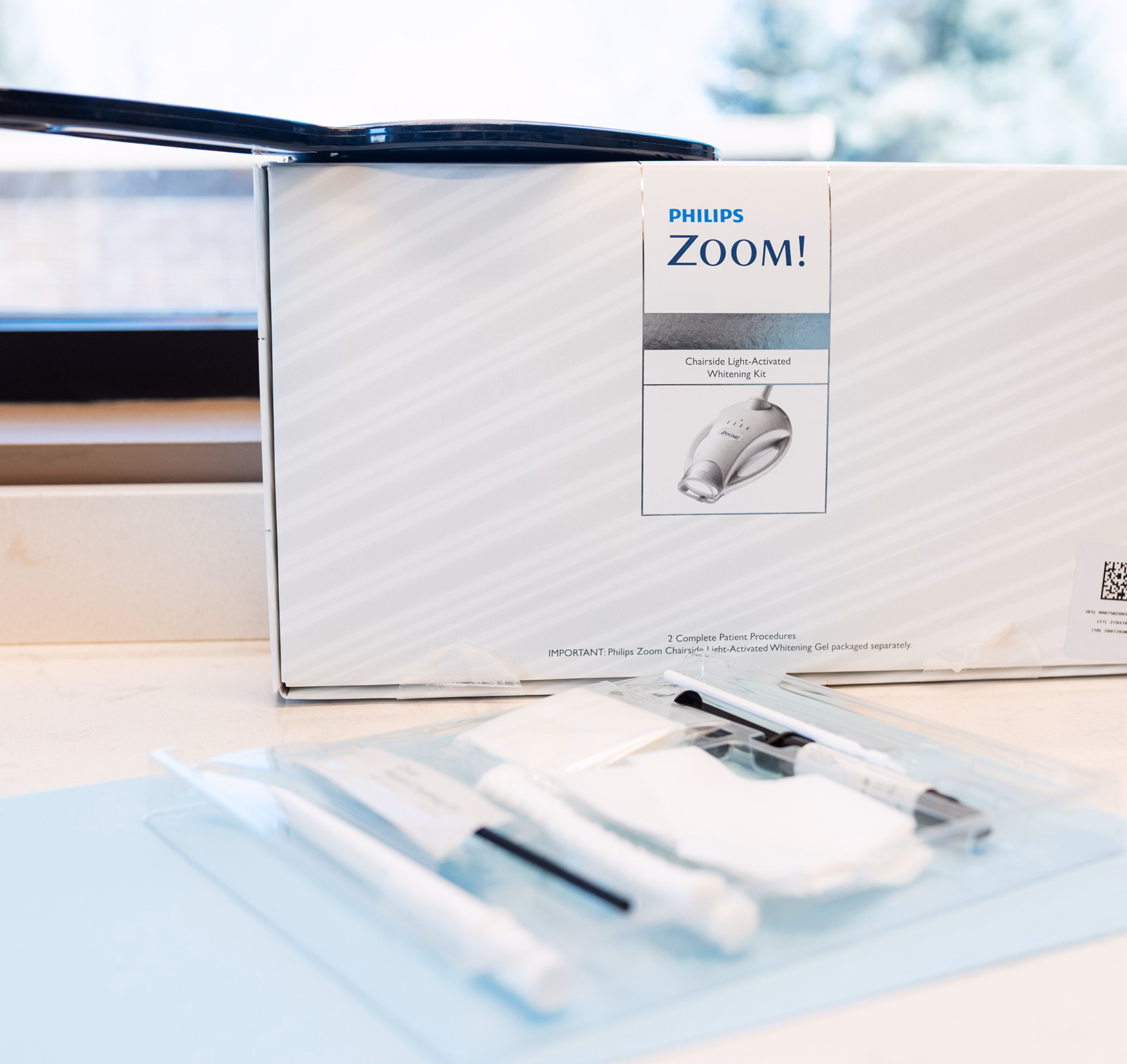 A Philips Zoom! chairside light-activated whitening kit box is on a countertop. In front, a clear tray holds dental tools and accessories. Bright and clinical setting.