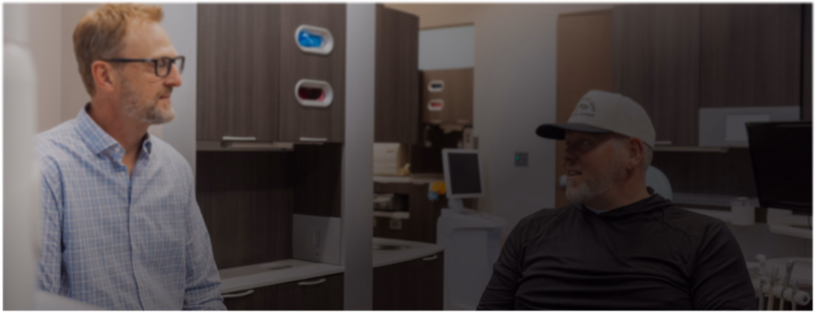 Two men are talking in a modern office setting with wood cabinets and computer screens. The atmosphere is casual and focused.