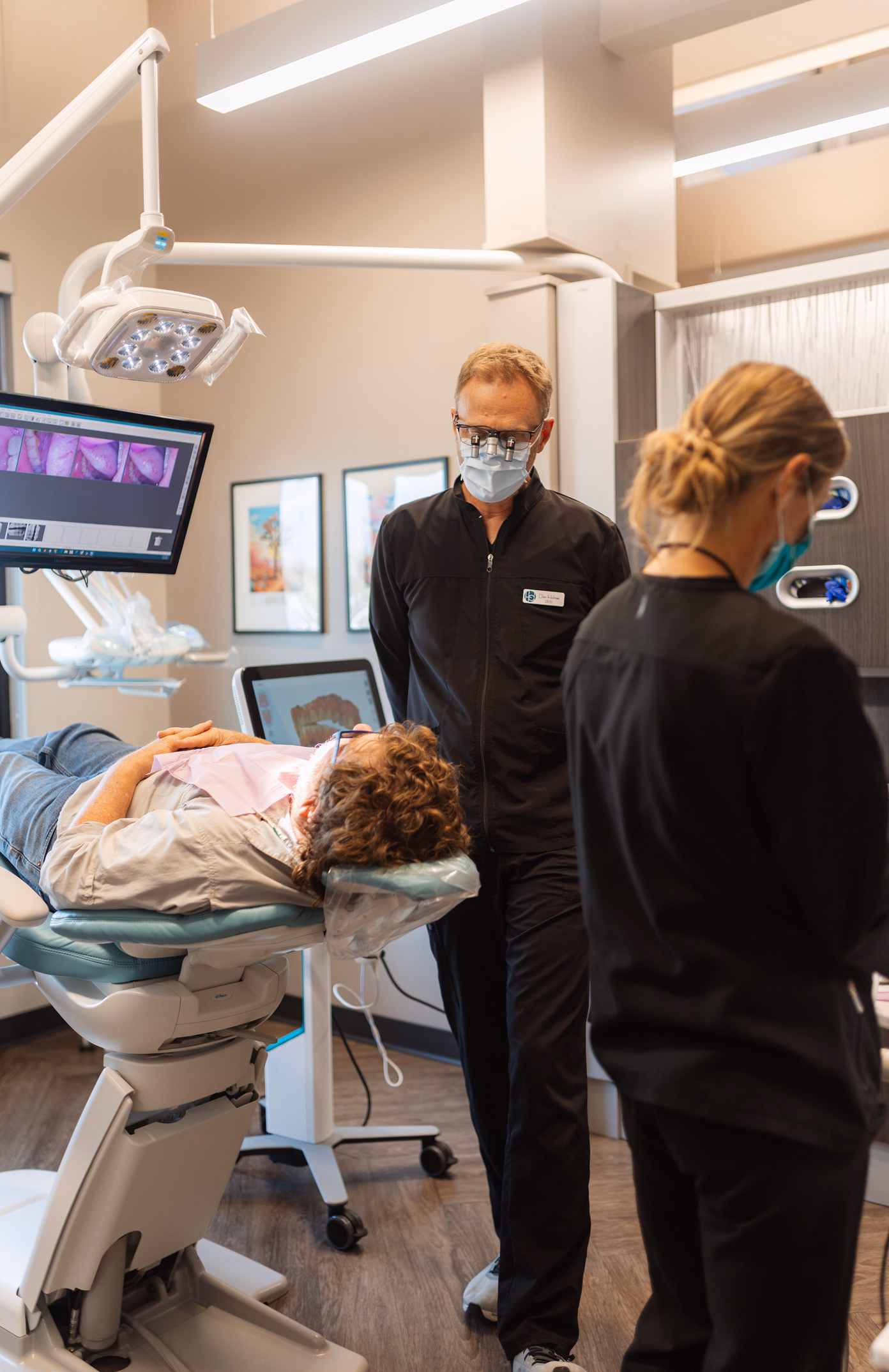 A dentist in black scrubs and a mask stands near a patient lying on a reclined dental chair. A screen displays dental images. The setting appears clinical and professional.