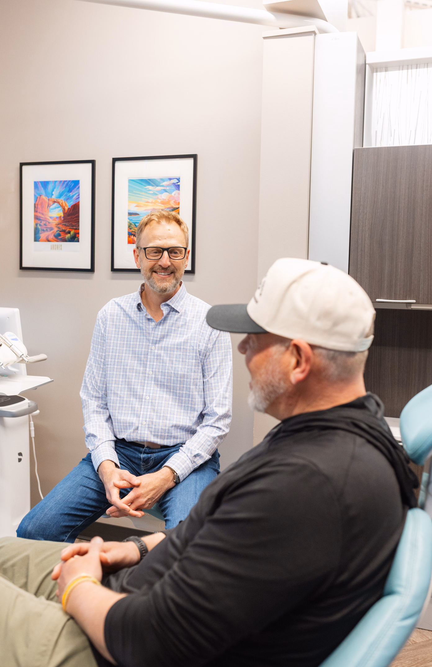 A dentist, wearing glasses and a plaid shirt, warmly converses with a patient seated in a dental chair. Colorful art decorates the office walls.