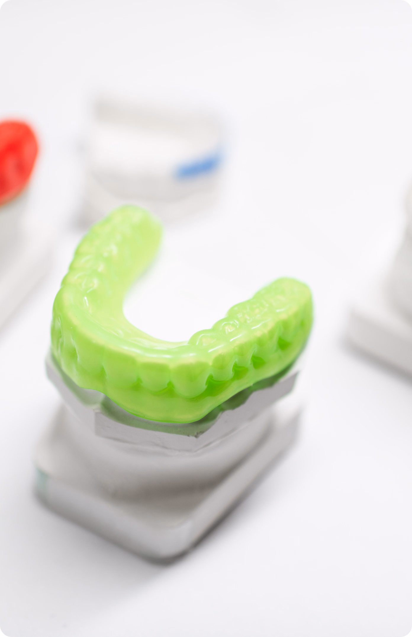 A close-up of a green dental mold on a white stand, with blurred red and blue molds in the background, conveying a neutral, clinical tone.