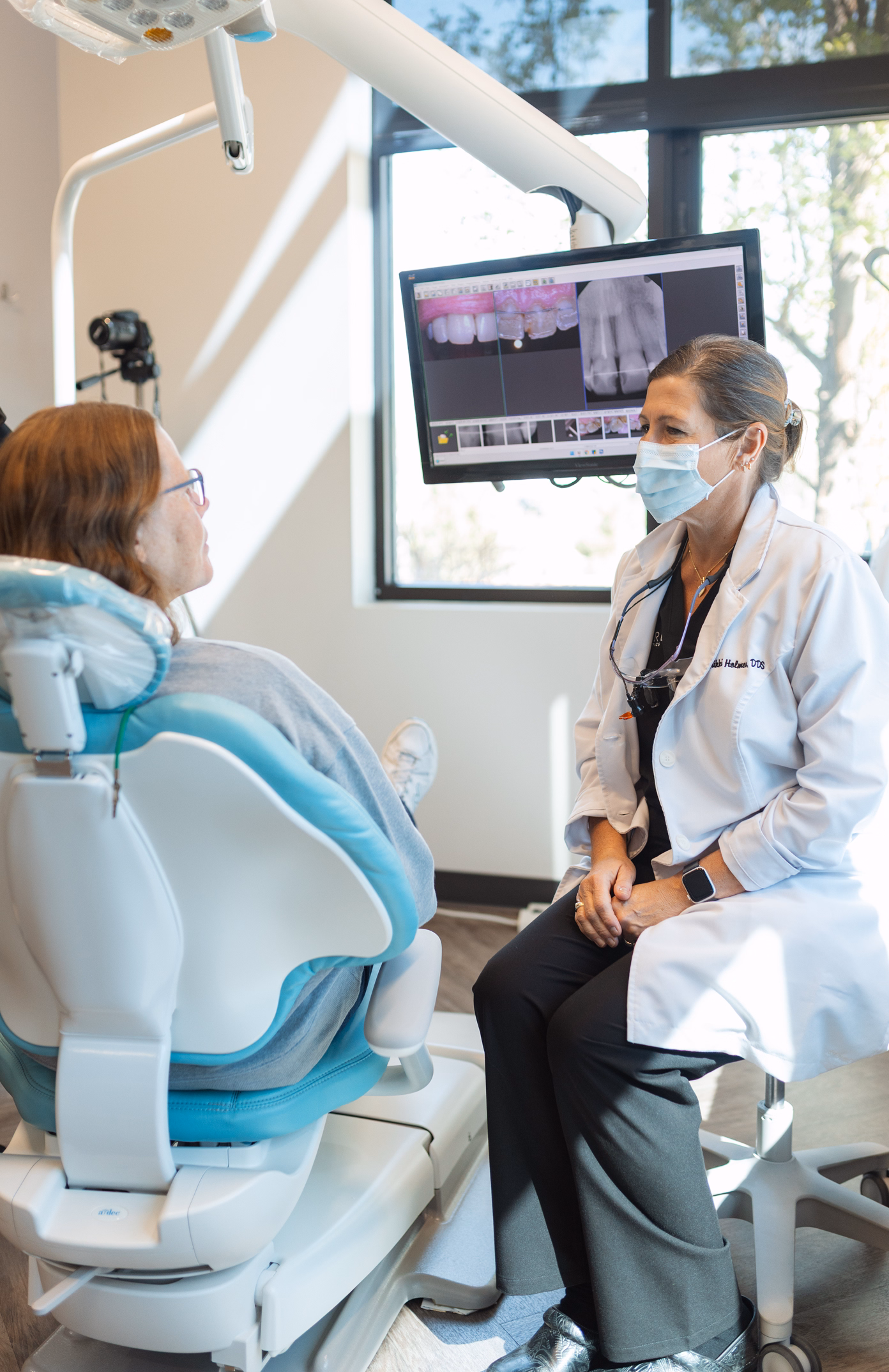 Dentist wearing a mask consults with a patient in a dental chair, with X-rays displayed on a screen. Bright, professional clinic setting.