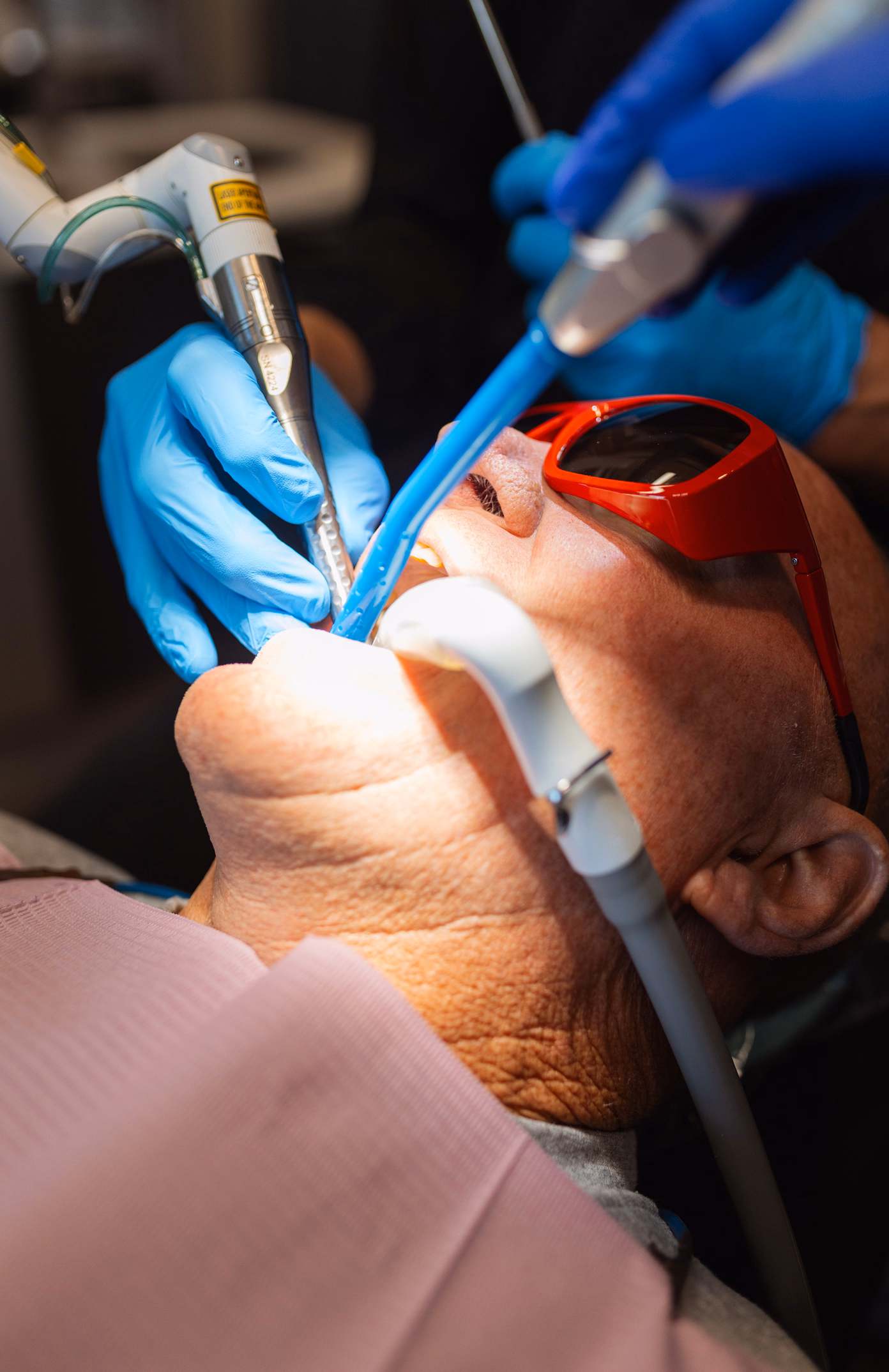Patient wearing red protective glasses and pink bib undergoing dental treatment. Dentist's gloved hands hold tools, creating a clinical atmosphere.
