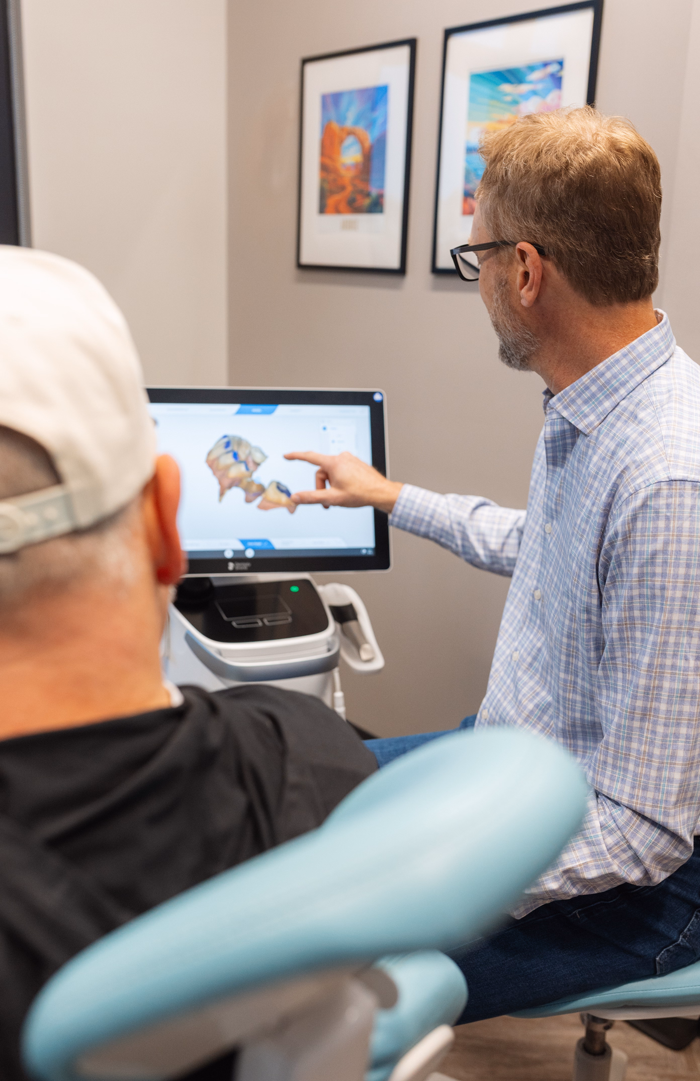 A dentist shows a 3D dental model on a screen to a patient sitting in a dental chair. Two colorful art pieces are on the wall, creating a calm atmosphere.