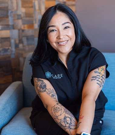 Savannah - Smiling woman with tattoos sits on a blue sofa. She wears a black shirt with a logo, against a warm-toned stone wall background, conveying a friendly tone.