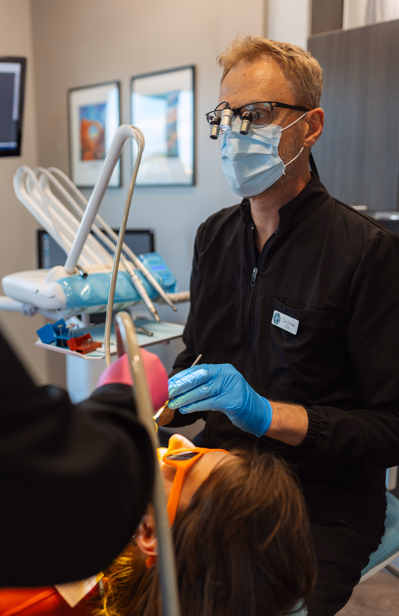 Dentist in a black coat and mask works on a patient's teeth in an office, wearing magnifying glasses. The patient wears orange protective glasses.