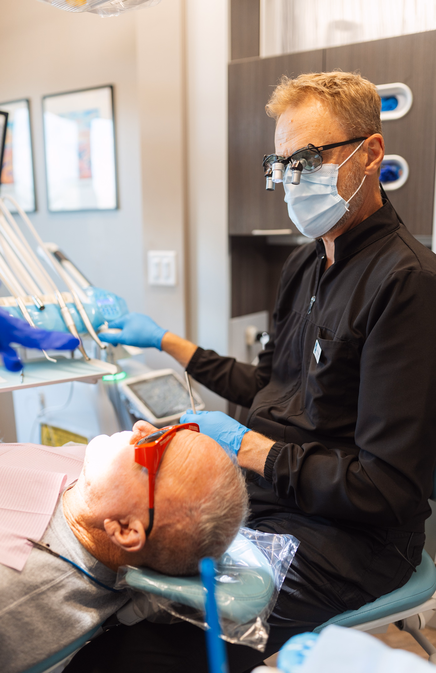 Dentist wearing magnifying glasses and a mask examines an elderly patient reclining in a dental chair. The patient wears protective glasses.