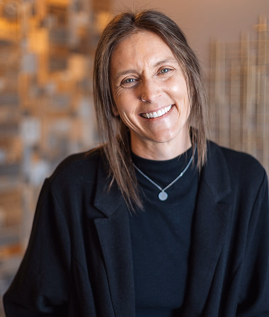 Betsy - Smiling in a black blazer and necklace, standing against a blurred, warm-toned background. The atmosphere is friendly and welcoming.