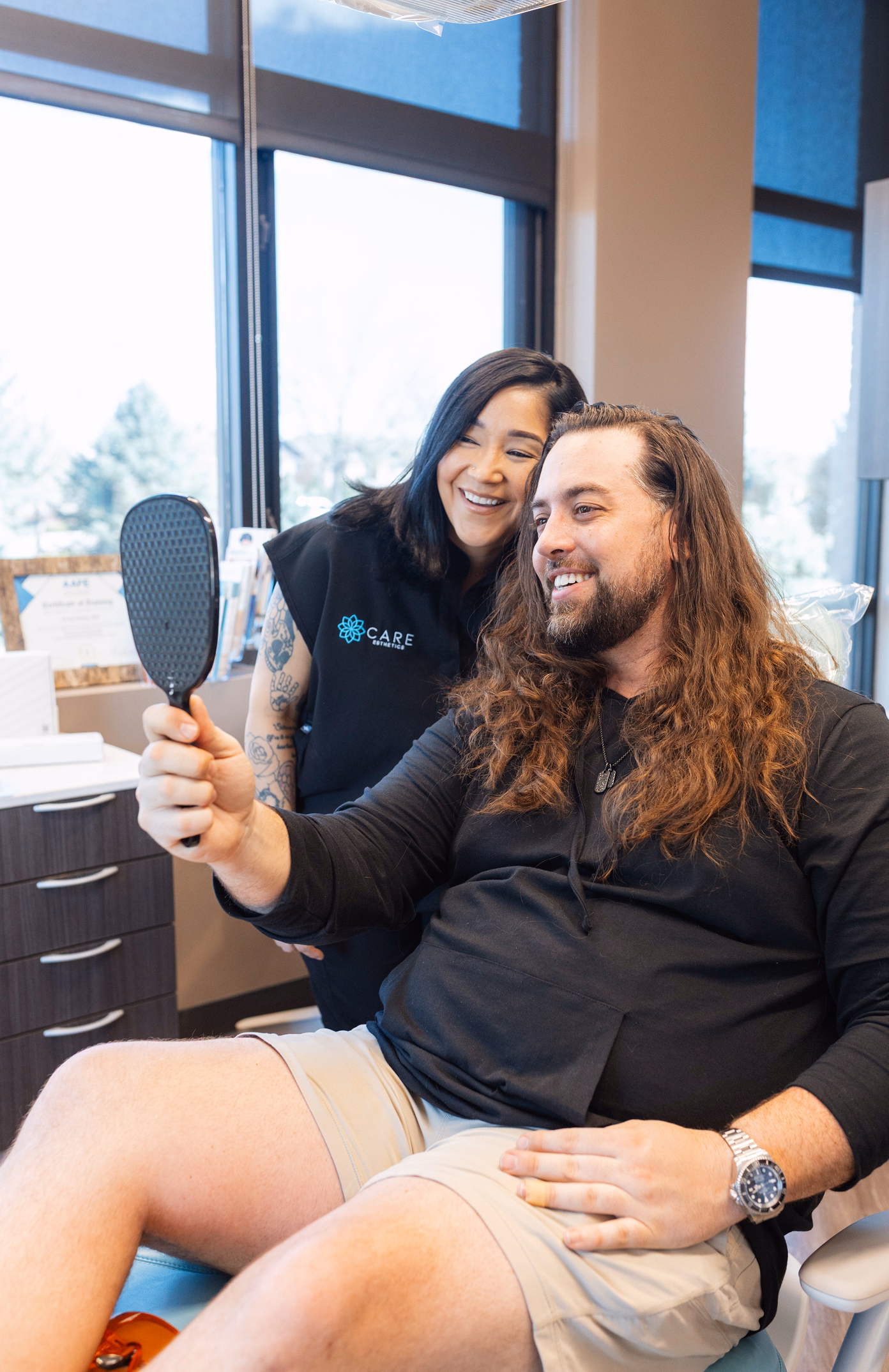 A man with long hair smiles while holding a mirror, sitting in a clinic. A smiling woman in a dental uniform stands beside him, creating a cheerful atmosphere.