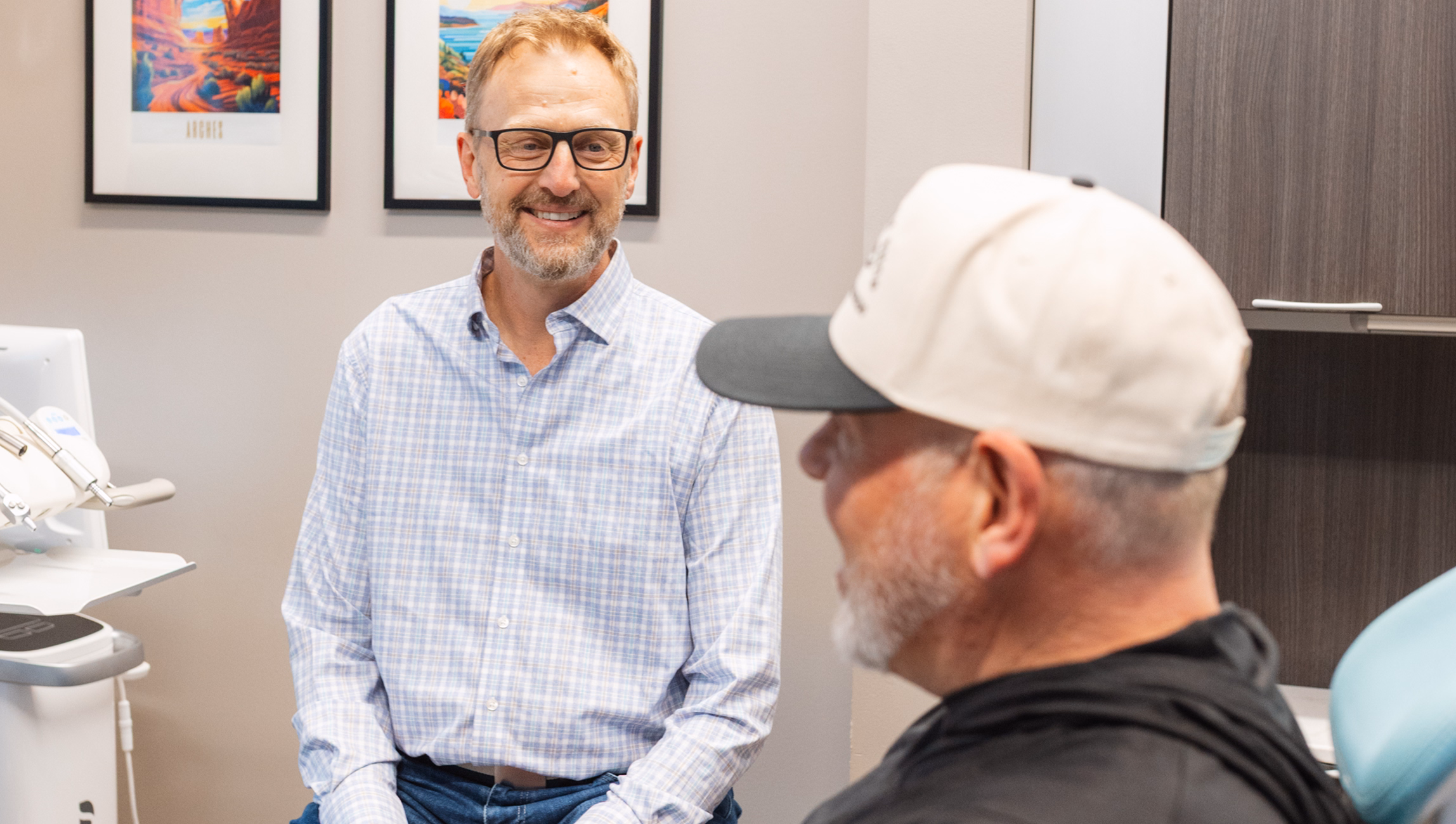 A man in glasses and a checked shirt smiles warmly at another man wearing a white cap in a bright office setting, conveying a friendly, professional atmosphere.