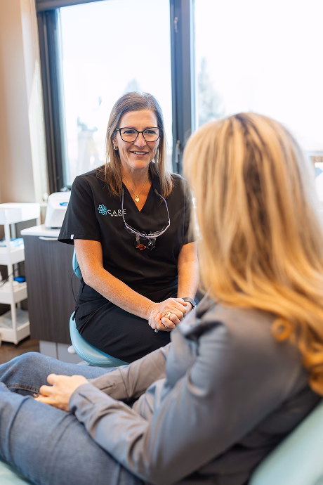 A dentist in black scrubs converses compassionately with a patient in a clinical setting. The atmosphere is calm and professional, with natural light.
