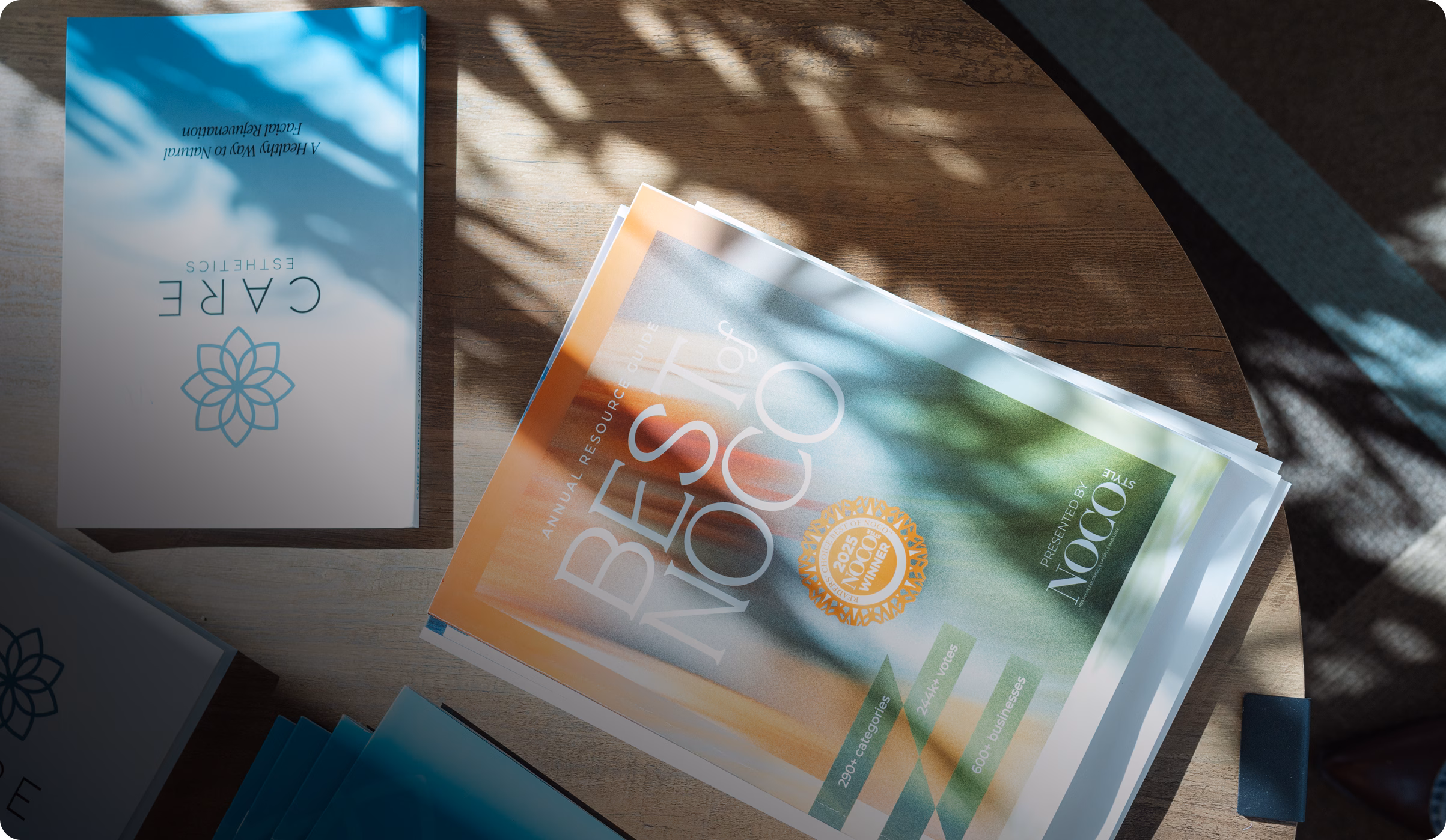 A wooden table with scattered magazines, including one reading "Best of NOCO" beside a "CARE" pamphlet. Leaf shadows create a serene atmosphere.