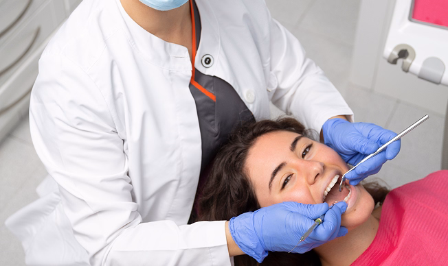 A dentist in a white coat and blue gloves examines a smiling patient's teeth. The patient is relaxed and wears a pink bib, conveying a calm atmosphere.