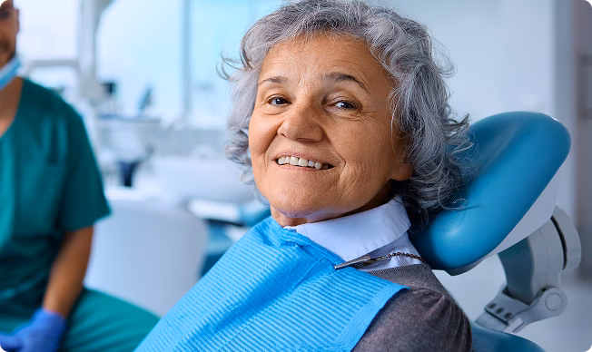 Smiling elderly woman with gray hair sits in a dental chair wearing a blue bib, dentist in scrubs partly visible. Bright, clean dental office.