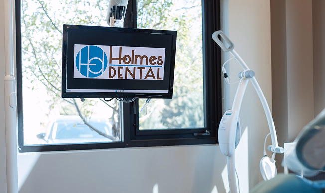 A dental office with a mounted screen displaying "Holmes Dental" logo. Bright sunlight filters through large windows, illuminating dental equipment.