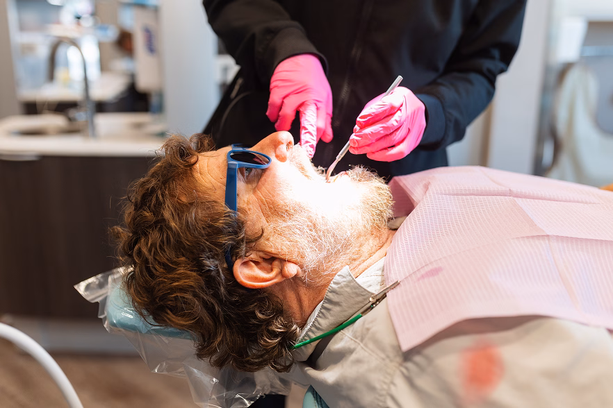 A dentist in black scrubs and pink gloves examines a patient wearing blue glasses in a dental chair, with a focus on oral care in a clinical setting.