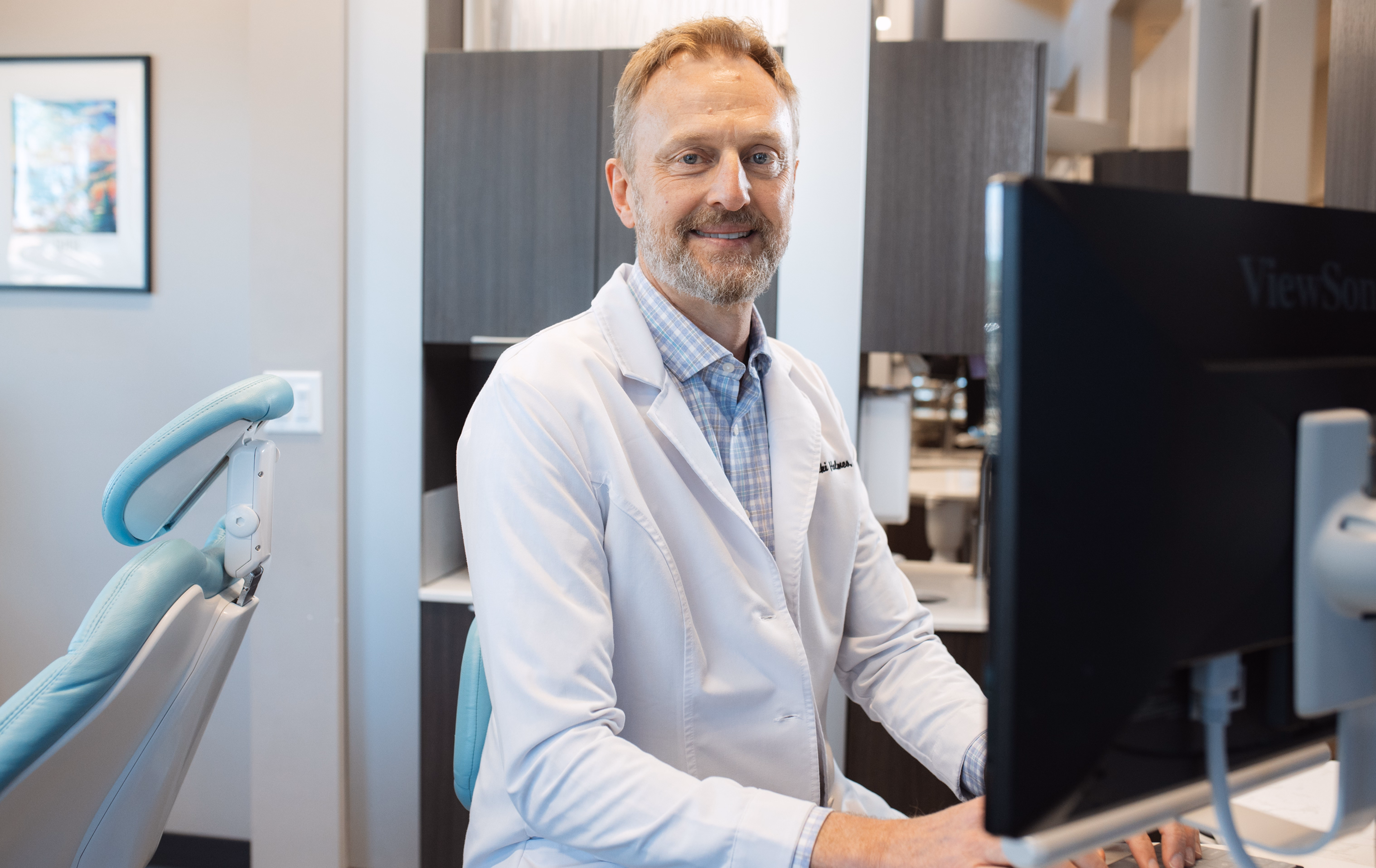 A middle-aged man in a white lab coat sits at a desk, smiling at a computer in a modern office setting, conveying a professional and approachable demeanor.