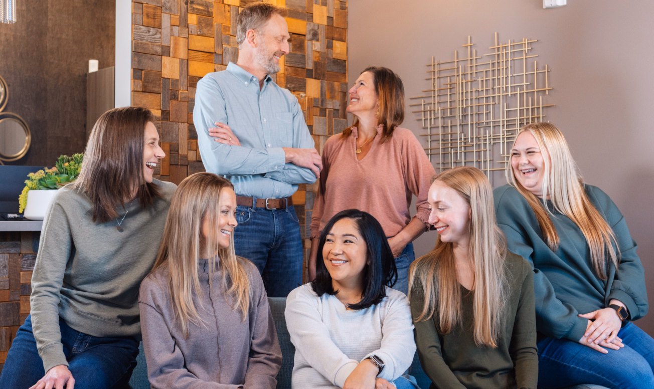 A group of seven people, six women and one man, are smiling and laughing indoors. The setting is cozy, with warm lighting and modern decor.