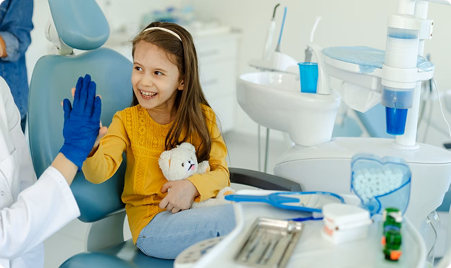 A young girl in a dental clinic, holding a teddy bear, smiles and high-fives a dentist in blue gloves. The mood is cheerful and reassuring.