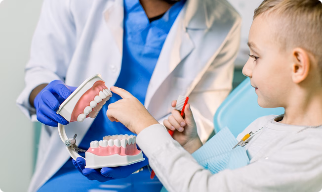 A child in a dental chair points to a tooth model held by a dentist wearing blue gloves and a white coat. The child holds a red toothbrush.