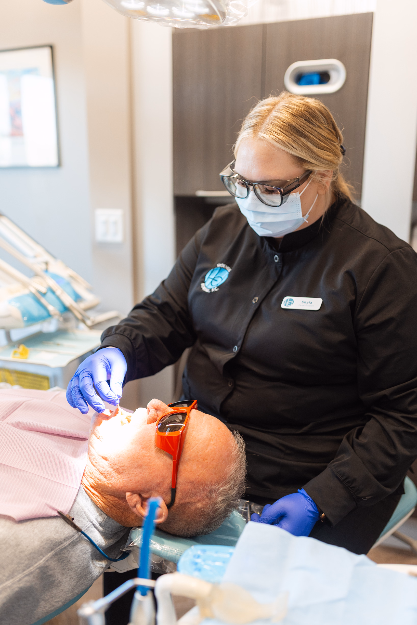 A dental professional wearing a mask and gloves examines a patient reclining in a dentist chair. The patient wears protective glasses, and the room is warmly lit.