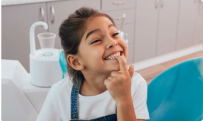 A smiling girl in a dental clinic points to her tooth, seated on a blue chair. She seems happy and playful, with a dental setup in the background.