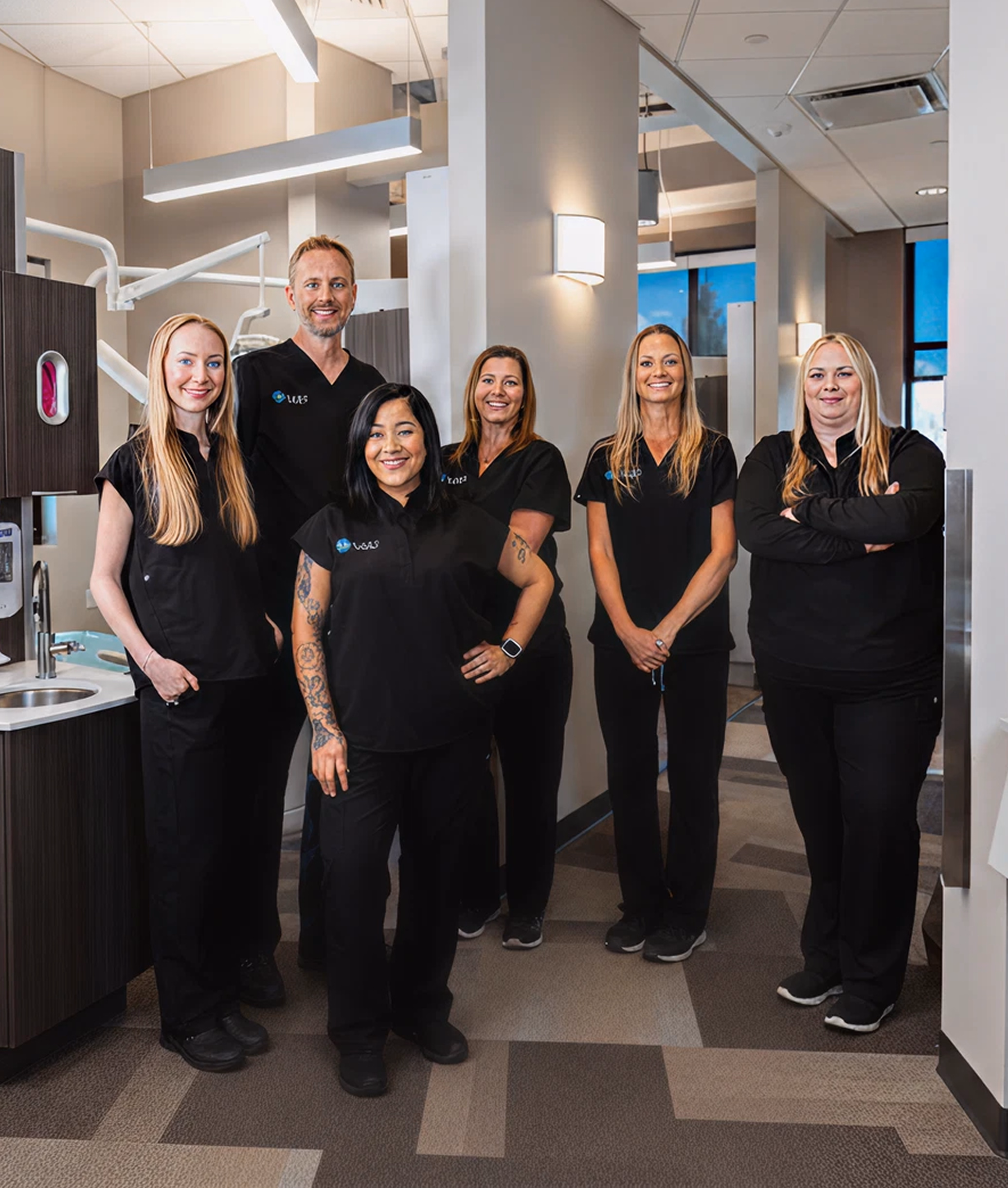 A group of six smiling individuals in black uniforms pose confidently in a bright, modern dental office, conveying a welcoming and professional atmosphere.